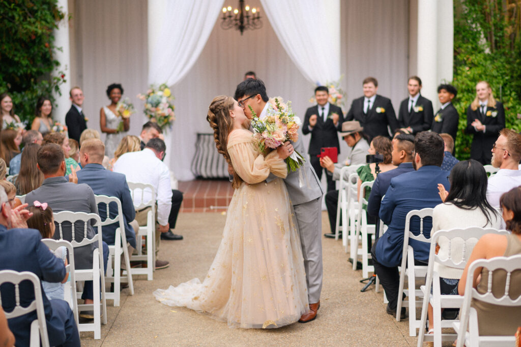 Wedding ceremony at Gray Gables Estate featuring a bride in a golden gown and groom sharing a kiss at the altar, surrounded by guests seated outdoors.