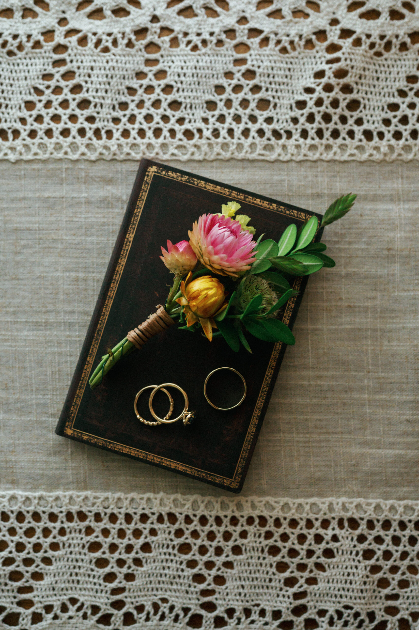 A small boutonniere with pink and yellow flowers is placed on an antique leather-bound book, alongside a pair of gold wedding rings and an engagement ring. The intricate lace tablecloth beneath adds a timeless, heirloom feel to the composition.