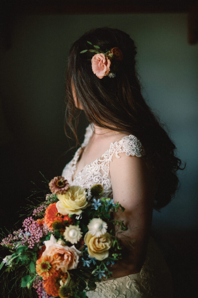 A close-up of the bride’s hair, styled in soft waves and adorned with a peach-colored flower. She holds her bouquet of lush, colorful blooms, the texture of her lace gown adding depth to the composition. The intimate lighting enhances the natural elegance of the moment.