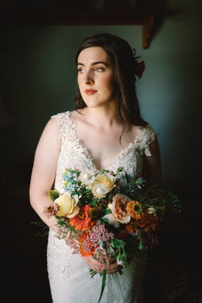 The bride stands in soft, directional light, holding a bouquet of wildflowers in warm, earthy tones. Her lace wedding gown features delicate floral details, and she gazes off with a quiet, contemplative expression. The moody lighting and natural textures create an intimate, timeless feel.