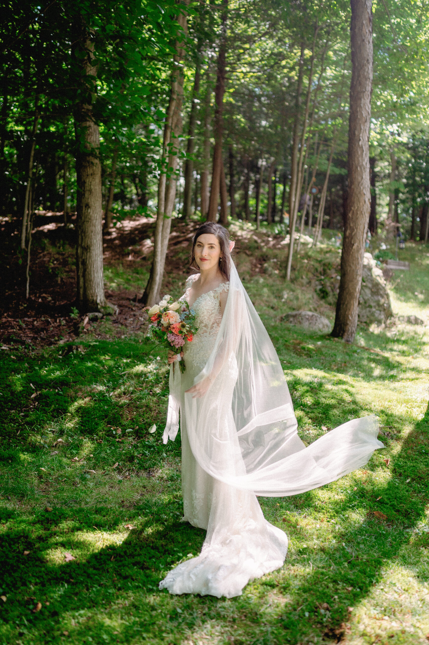 A bride stands in a sunlit forest clearing, her lace gown flowing elegantly as her veil catches the breeze. She holds a bouquet of warm-toned flowers, her expression calm and content. Dappled sunlight filters through the trees, casting soft shadows on the mossy ground, creating a serene and natural atmosphere.