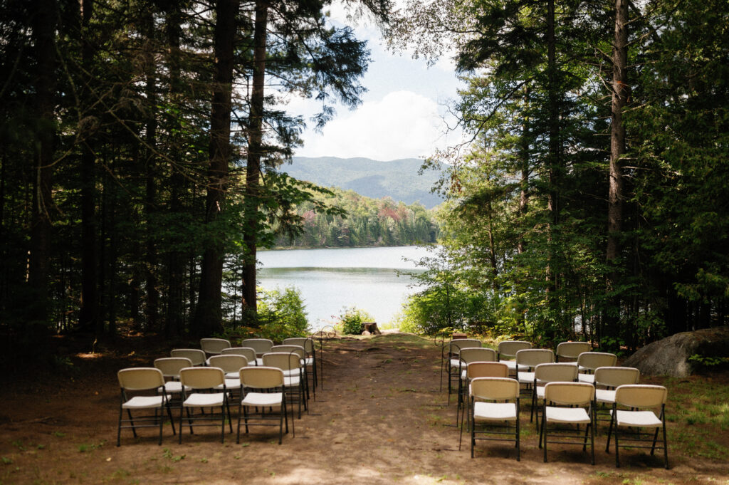 A serene lakeside ceremony setup at Heart Lake – Rows of simple folding chairs face the water, framed by tall pine trees, creating an intimate outdoor wedding setting with the Adirondack mountains in the distance.