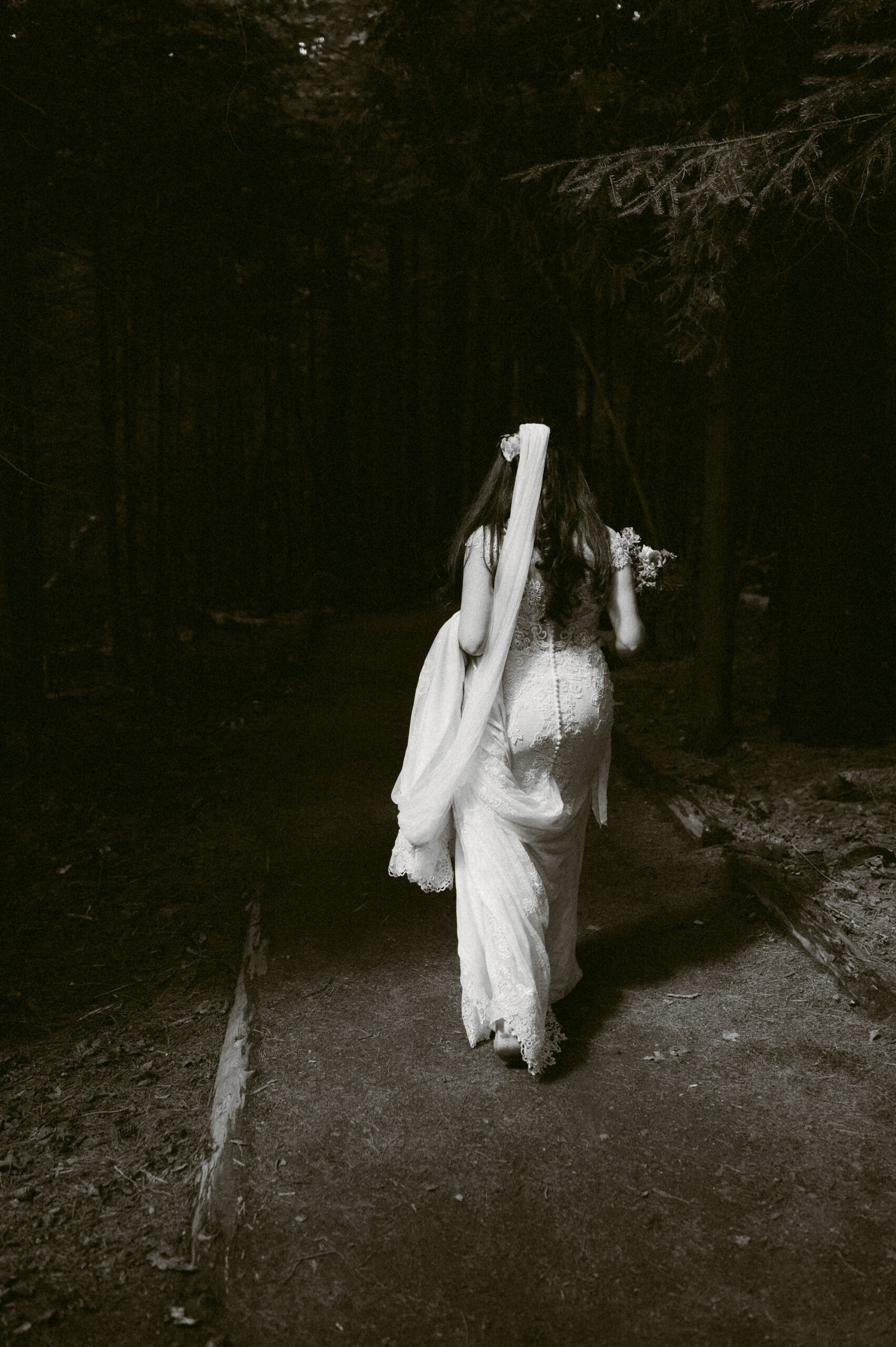 A bride walks along a quiet forest path, her veil trailing behind her as she approaches her groom for their first look. The black-and-white treatment enhances the timeless, intimate feel of the moment.