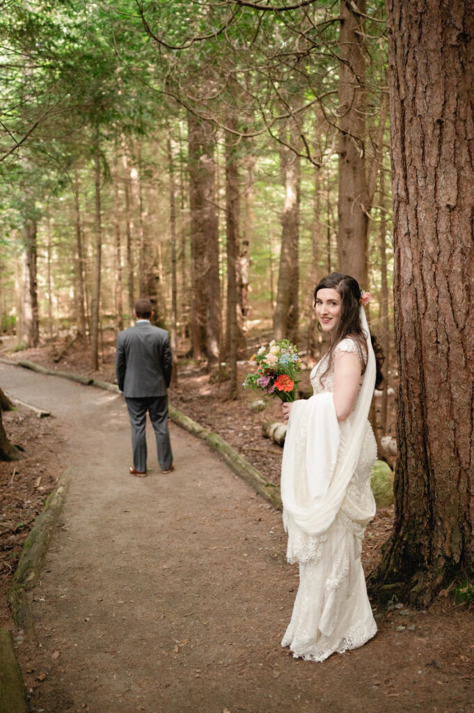 The bride pauses behind a tree, a soft smile on her face as she holds her bouquet and looks toward her groom, who stands further down the path with his back turned, waiting for their first look. The surrounding trees frame the scene beautifully.