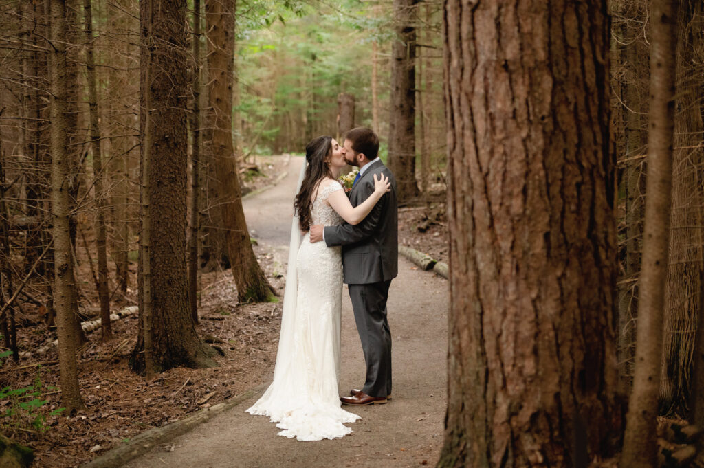 The couple shares a kiss in the middle of the trail, surrounded by towering trees. The composition draws the eye down the winding path, creating a sense of depth and intimacy.