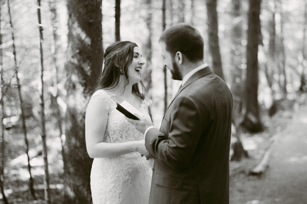 In a black-and-white image, the bride bursts into laughter as the groom reads his vows, their emotions unfolding naturally in the peaceful surroundings. The candid moment captures the joy and authenticity of their connection.