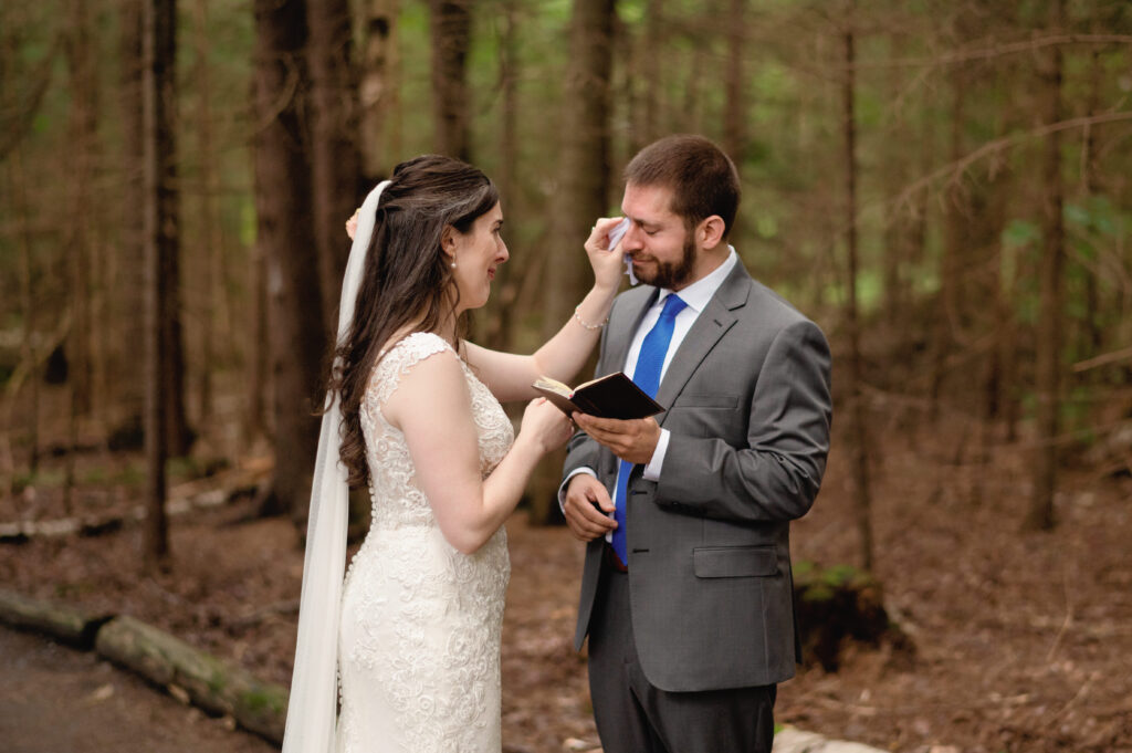 The bride gently wipes away a tear from the groom’s face as he holds a small notebook, reading his private vows. The quiet forest setting adds to the intimacy of the moment, highlighting their deep connection.