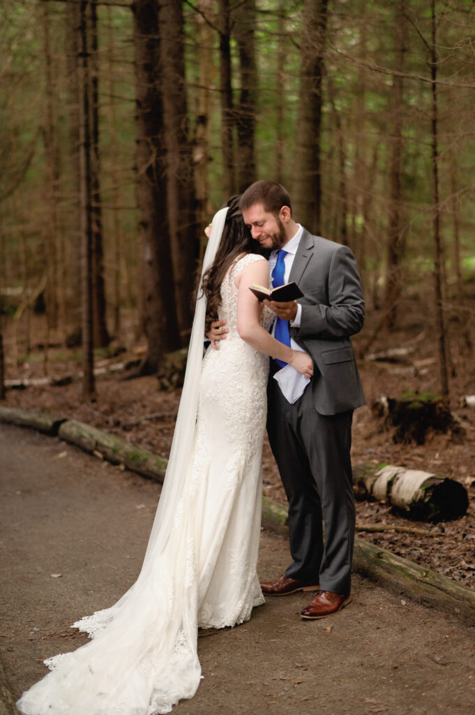 The groom leans into the bride, holding his vow book close as they embrace after sharing their words. The moment feels deeply personal, framed by the towering trees around them.