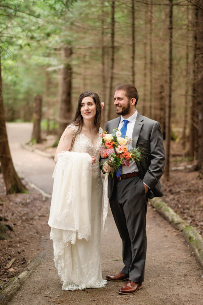 Standing together on a wooded path, the couple takes a moment to reflect after exchanging their vows. The bride holds her lace gown and bouquet, while the groom, dressed in a gray suit and blue tie, smiles warmly.