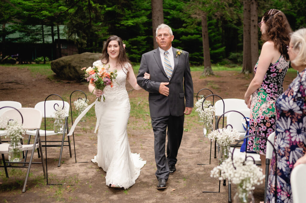 Bride walking down the aisle with her father during her Adirondack Loj wedding – She smiles while holding a vibrant bouquet of wildflowers as guests watch from their seats, surrounded by the peaceful forest setting.