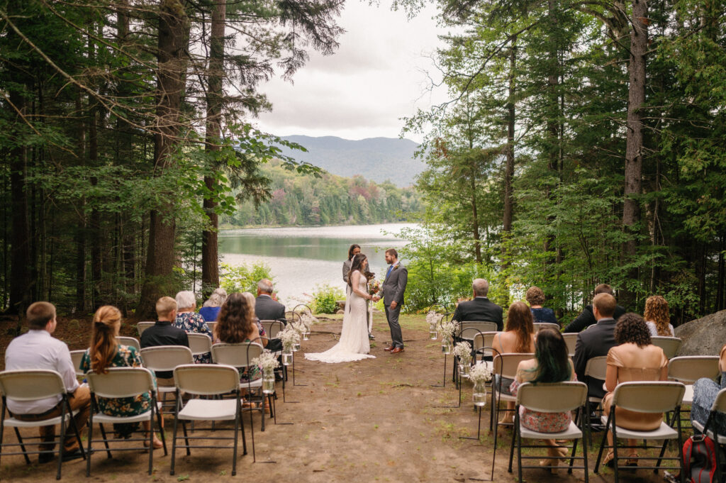 An intimate wedding ceremony at Heart Lake during their Adirondack Loj wedding – The couple stands together in front of their officiant, exchanging vows with the lake shimmering behind them as family and friends watch.