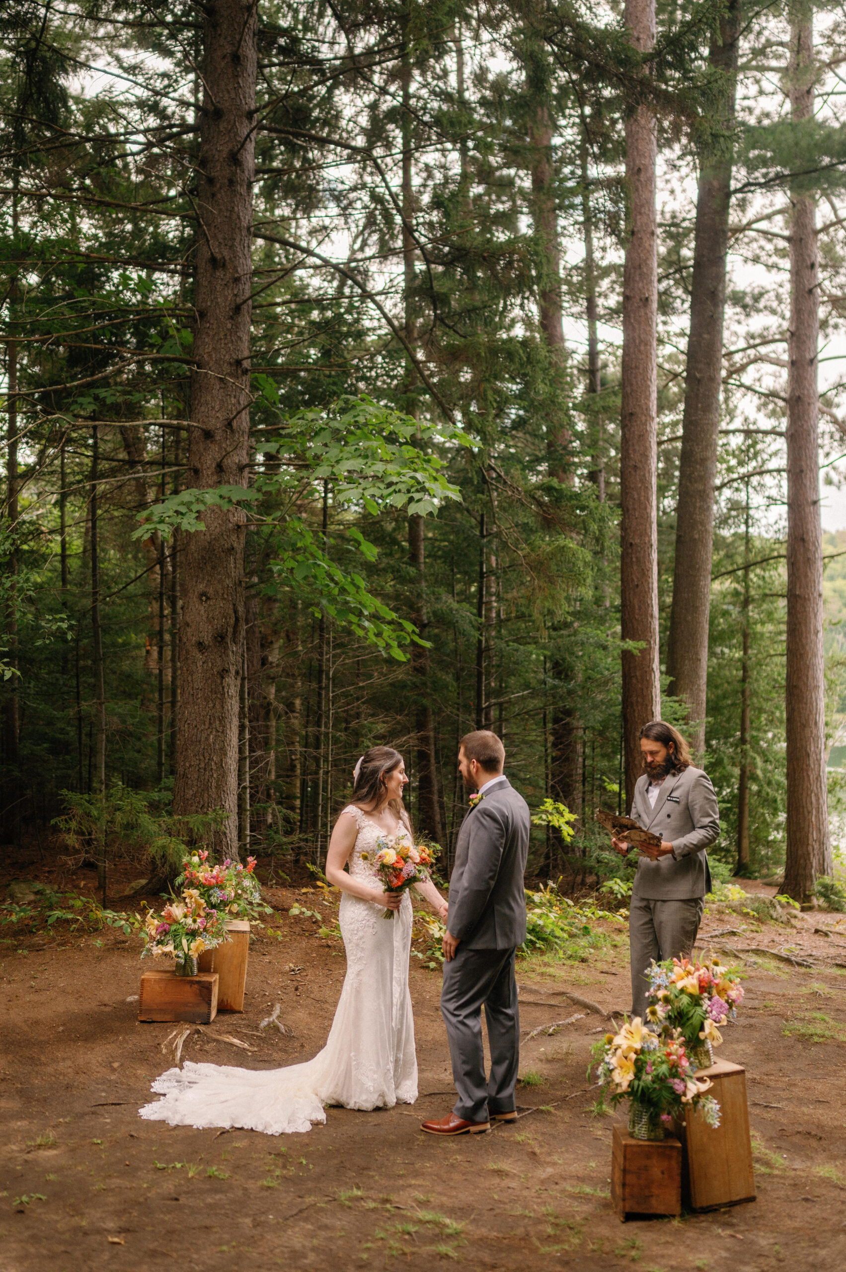 Bride and groom standing together in a wooded ceremony space during their Adirondack Loj wedding  – The officiant, dressed in a gray suit, reads from a book as the couple smiles at each other, surrounded by rustic floral arrangements and towering trees.