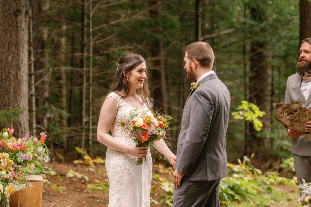 A quiet moment between the couple during the ceremony at their Adirondack Loj wedding – The bride, holding a colorful bouquet, gazes at the groom with soft eyes while the officiant speaks, their connection evident in the peaceful forest setting.