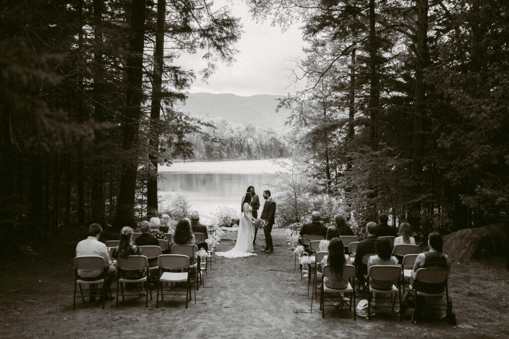 Black and white image of an intimate outdoor wedding ceremony at Heart Lake – Guests sit in rows of chairs nestled among tall trees as Eric and Martha stand before their officiant, exchanging vows with the lake and mountains as their backdrop.