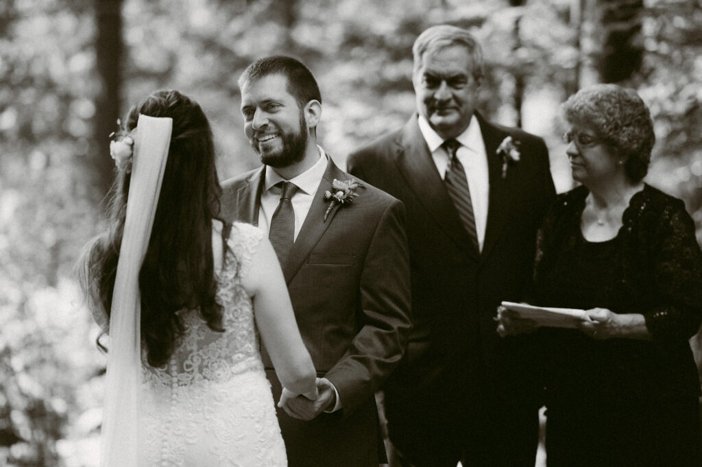 Black and white image of groom smiling during vows – Eric gazes at Martha with a loving smile as they exchange vows, while two family members stand behind them, watching the moment unfold.