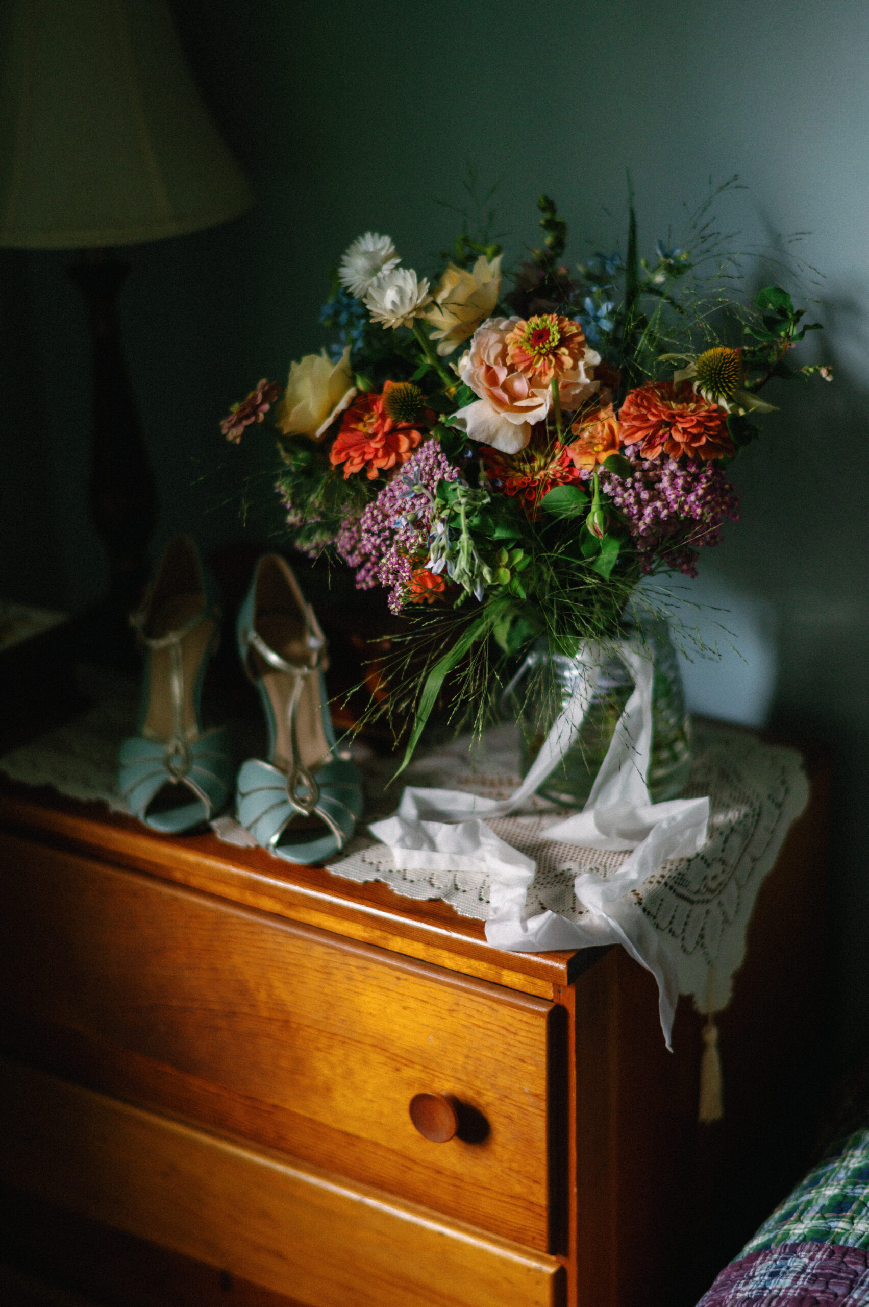 A vibrant wedding bouquet with orange, peach, and blue flowers sits in a glass vase on a wooden nightstand, accompanied by a pair of vintage-inspired teal heels and a delicate white ribbon. Soft, moody lighting highlights the rich textures and colors.