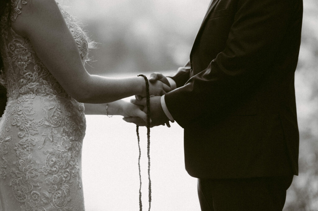 Black and white close-up of a handfasting ceremony during their Adirondack Loj wedding – The couple’s hands are wrapped in a braided cord as part of a symbolic handfasting ritual, with the soft glow of the lake in the background.