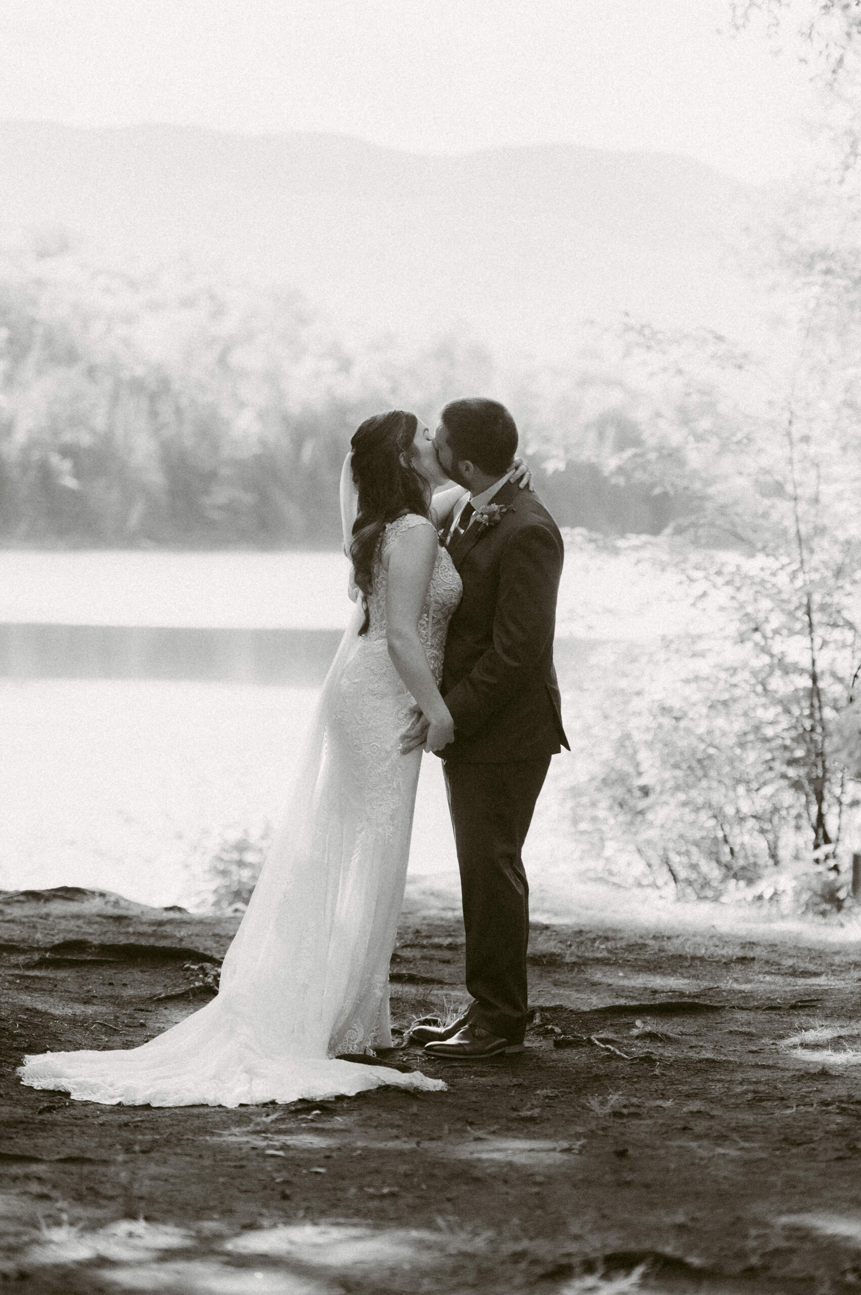 Black and white image of bride and groom sharing a kiss by Heart Lake during their Adirondack Loj wedding – Eric and Martha share a kiss near the water’s edge, with the soft silhouette of the Adirondack Mountains in the background, creating a timeless and romantic moment.