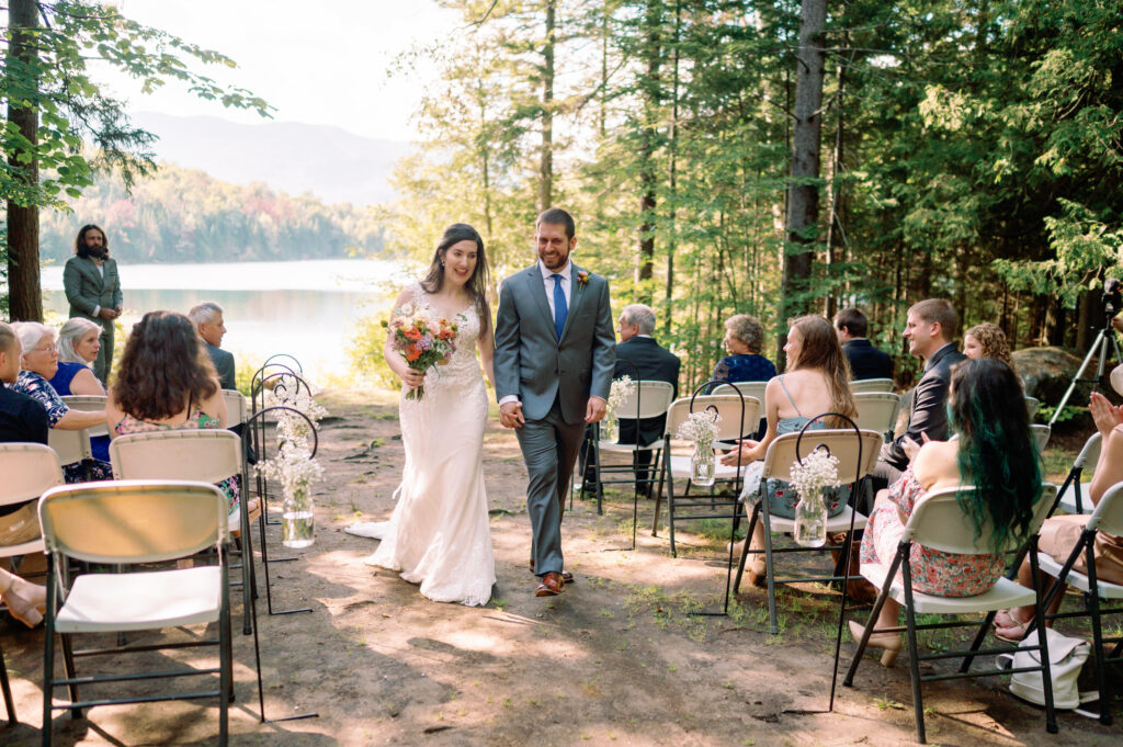 The newlyweds walk down the aisle together after their outdoor ceremony at Heart Lake, smiling as guests seated on white chairs watch and clap. The Adirondack mountains and the lake shimmer in the background, framed by towering trees. The bride’s flowing lace dress and bouquet contrast beautifully against the natural setting.