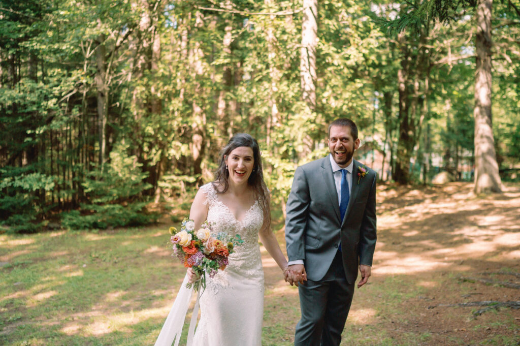 A joyful bride and groom walk hand in hand across a sunlit clearing in the forest, beaming with happiness after their ceremony. The bride holds a colorful bouquet, and the groom wears a gray suit with a blue tie. Lush green trees surround them, casting dappled light on the scene.