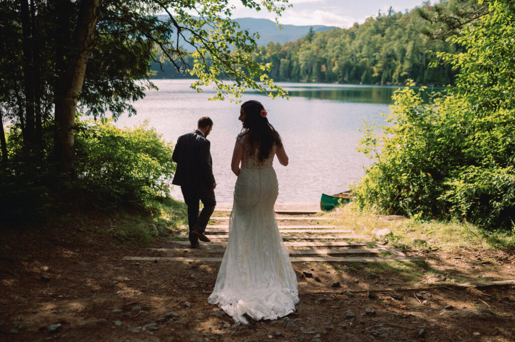 Bride and groom walking down the forested path toward Heart Lake during their Adirondack Loj wedding – The bride in her flowing lace gown follows the groom down a wooden staircase leading to the water, where their canoe awaits.