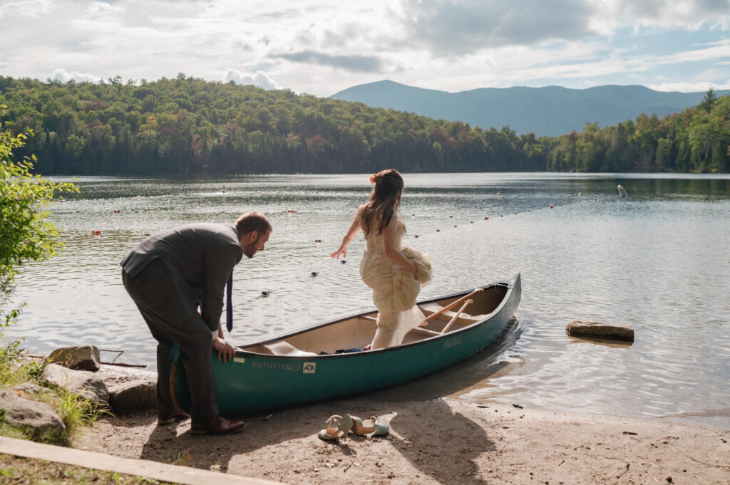 Bride stepping into a canoe while the groom steadies it on the shore of Heart Lake during their Adirondack Loj wedding – The groom, in a gray suit, holds the canoe steady as the bride lifts her dress and steps in, with her green heels left on the sandy shoreline.