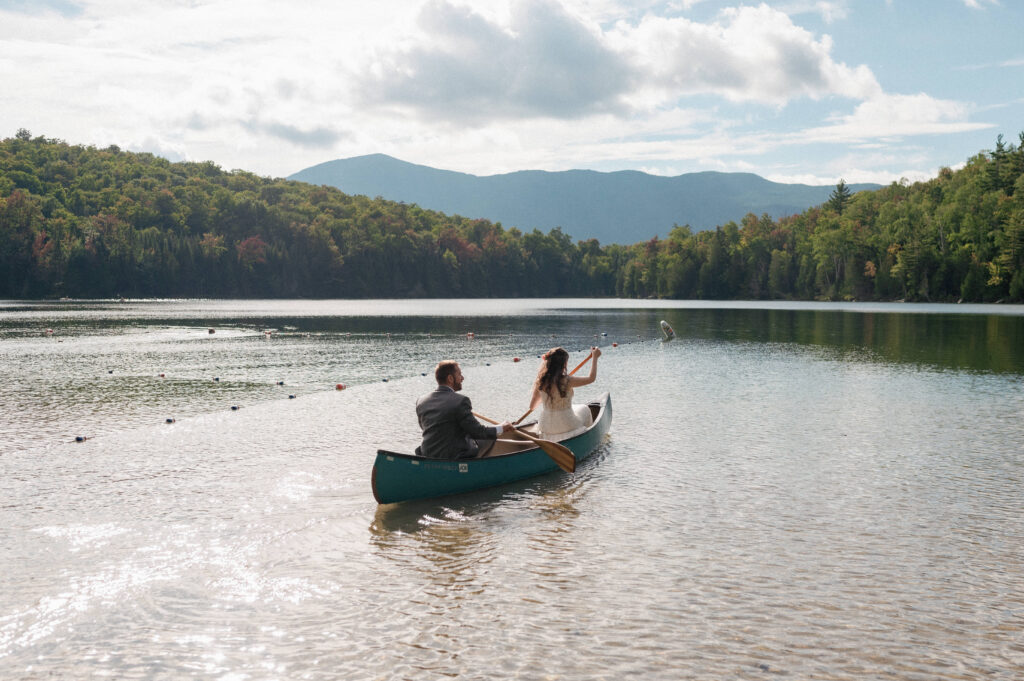 Bride and groom paddling across Heart Lake in a green canoe during their Adirondack Loj wedding – The couple glides across the water, surrounded by the lush forest and mountain views of the Adirondacks, as the bride raises her paddle in a moment of joy.