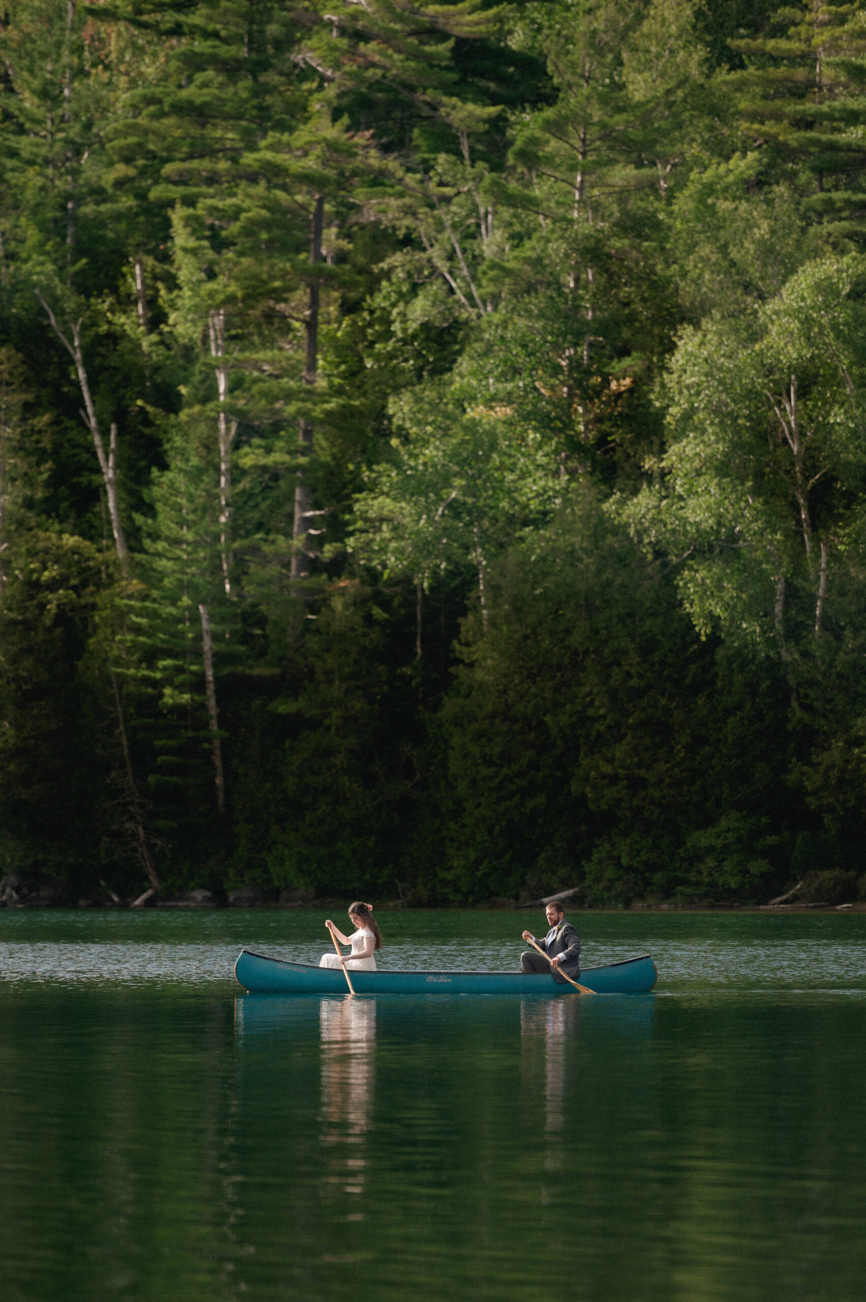 Bride and groom canoeing together on the still waters of Heart Lake during their Adirondack Loj wedding– A distant view of the couple paddling side by side, framed by the deep green trees reflecting on the water’s surface.