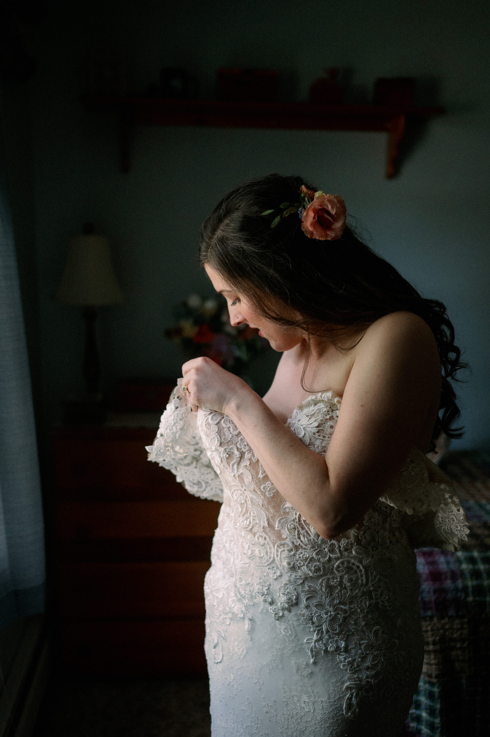 A bride adjusts the lace sleeve of her wedding gown as she gets ready in a softly lit room. Her dark hair cascades in loose waves, adorned with a delicate peach-colored flower. The intimate setting, with a wooden dresser and a bouquet in the background, adds warmth and nostalgia to the moment.