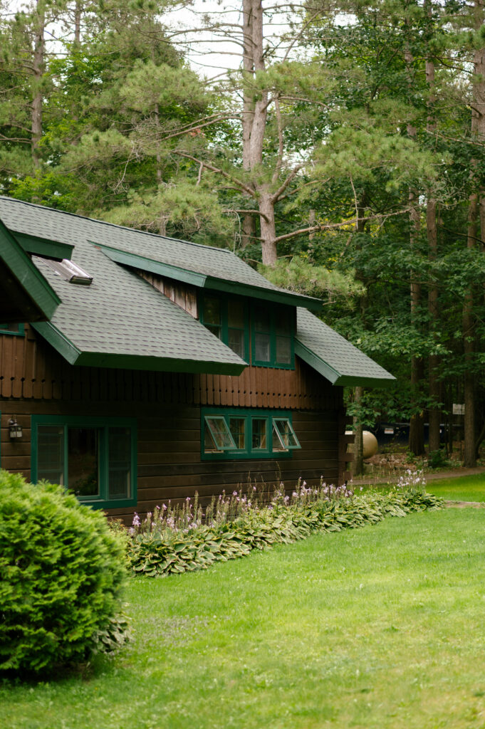 A cozy, dark brown Adirondack lodge with green window frames sits nestled among towering pine trees. The lush lawn and well-tended garden beds create a peaceful, inviting atmosphere.