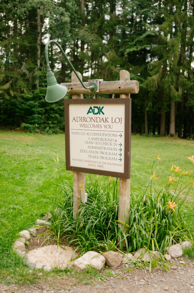 A rustic wooden sign welcomes visitors to the Adirondak Loj, with a vintage green lamp hanging above it. The sign provides directions to accommodations, trails, and educational programs, surrounded by lush greenery and blooming flowers.