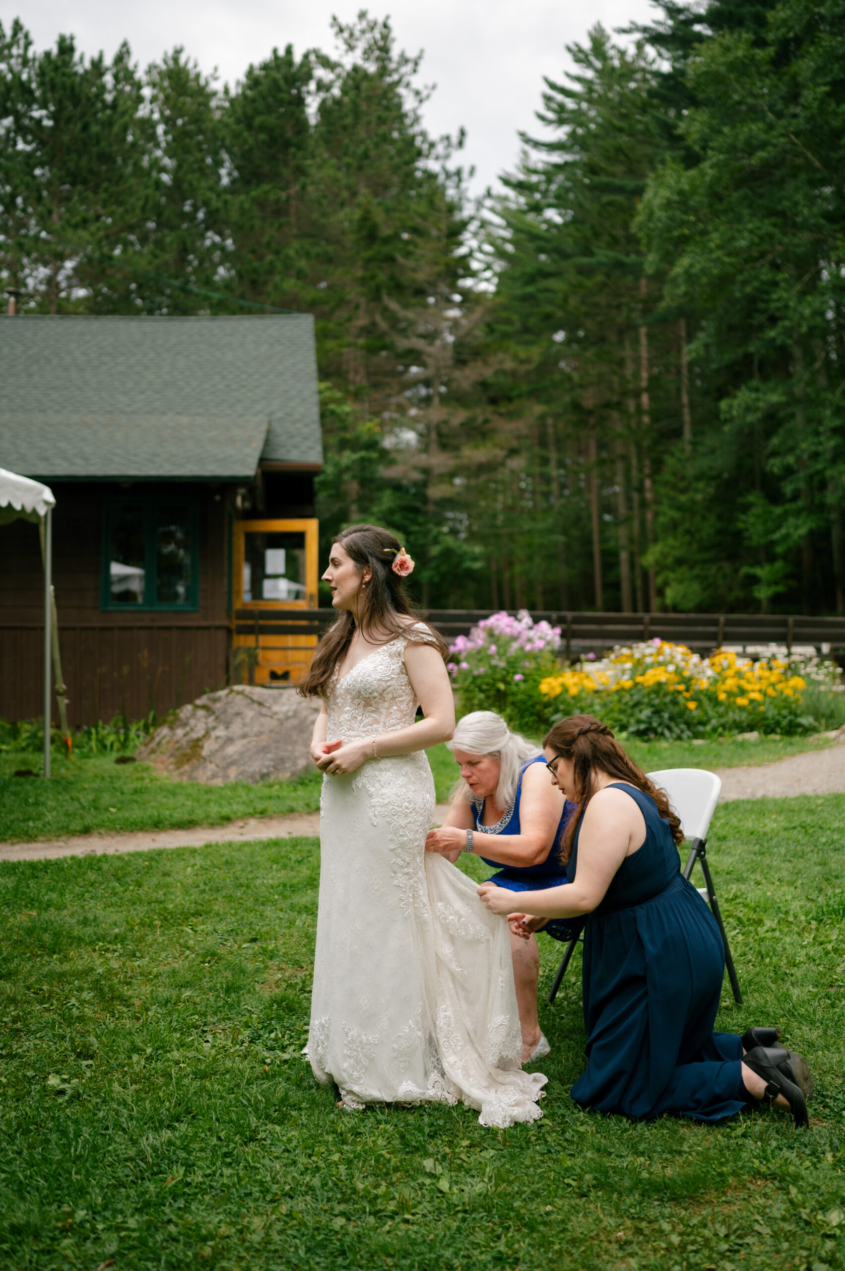 The bride stands on a green lawn while two women, one older and one younger, adjust the train of her lace wedding gown. Behind them, the rustic lodge and vibrant summer flowers add to the serene, outdoor setting.