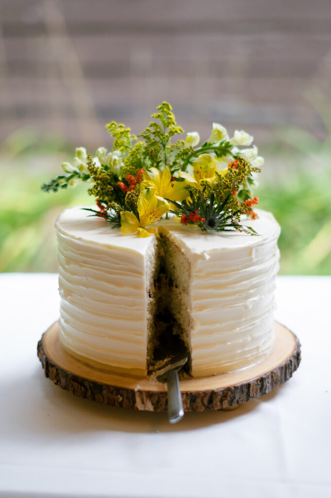 Rustic wedding cake with floral decorations and a slice missing – A round, white-frosted wedding cake with textured icing and fresh yellow and orange wildflowers sits on a wooden slab. A large slice is missing, revealing a blueberry-filled interior. A cake knife rests inside the cut section. The background is softly blurred, with hints of greenery.