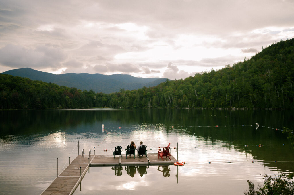 A dock extends into a calm lake at sunset, with Adirondack chairs arranged at the end. A couple sits facing the water, enjoying the view of mountains in the distance under a moody sky.