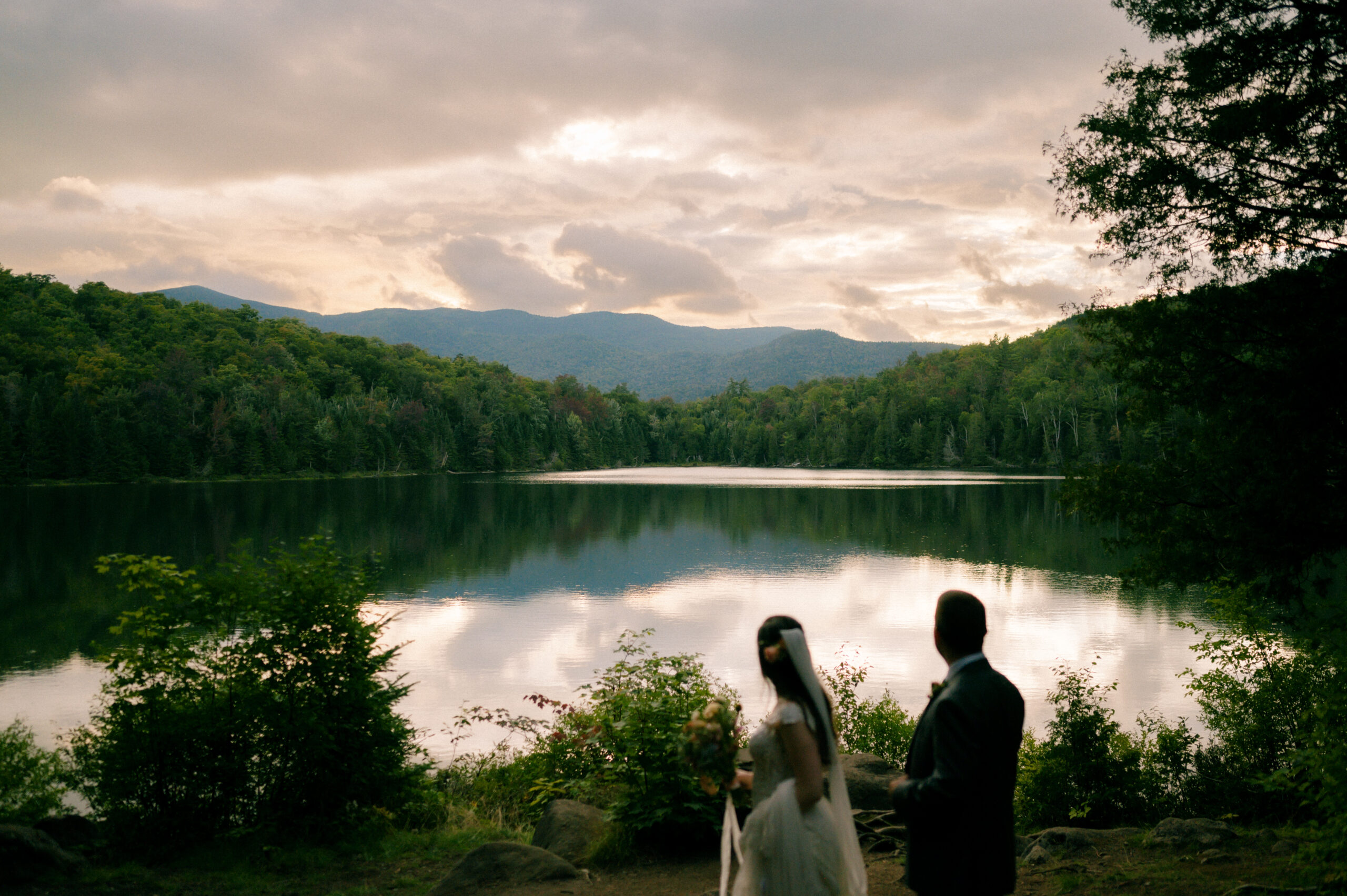 A bride and groom stand together at the edge of a tranquil lake, gazing at the reflection of the Adirondack Mountains beneath a cloudy sunset sky. The bride holds a bouquet, her veil gently draping behind her, while the groom, dressed in a suit, looks toward the water. Lush greenery frames the scene, creating a peaceful and intimate moment in nature.