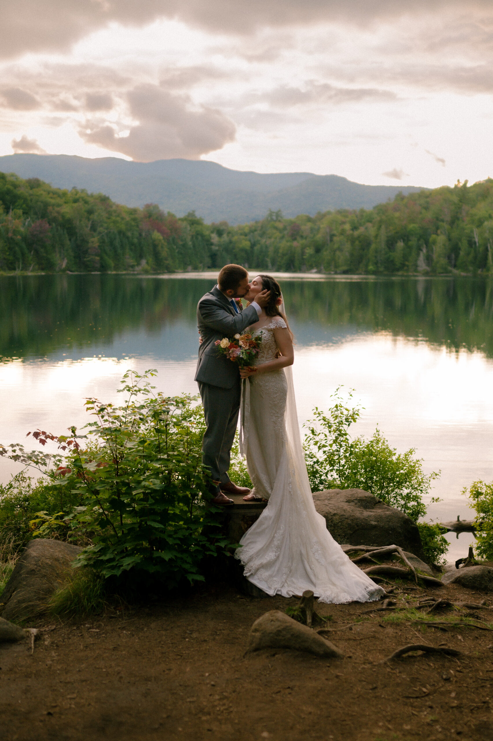 A bride and groom steal a kiss at the water’s edge as dusk settles over the mountains. Her veil spills over the rocks, his hand resting on her cheek, the lake reflecting the last colors of the day in a quiet, fleeting moment.