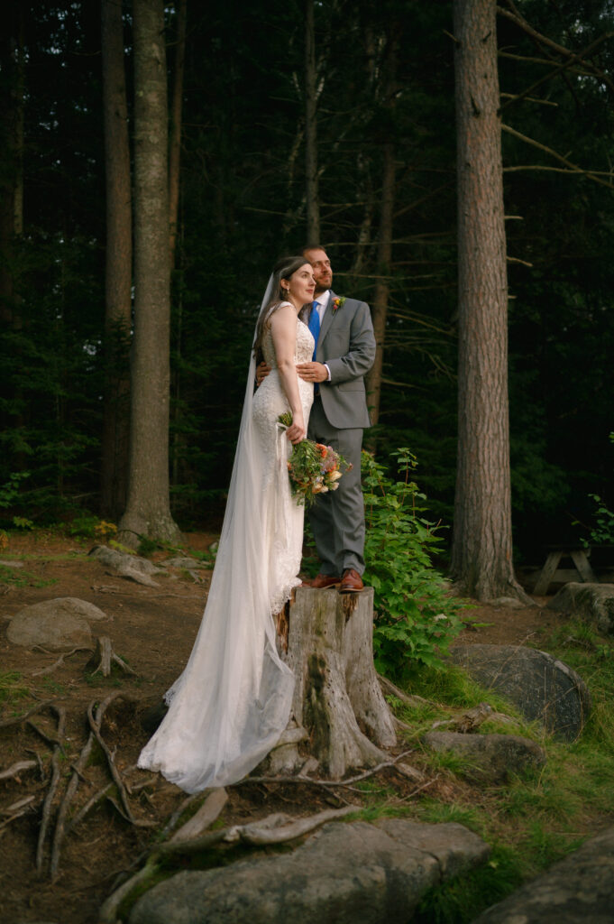 Bride and groom standing on a rugged tree stump, framed by towering pines. The golden hour glow catches the edge of the bride’s flowing veil as she holds a bouquet, her groom’s arm wrapped around her in a quiet, steady embrace.