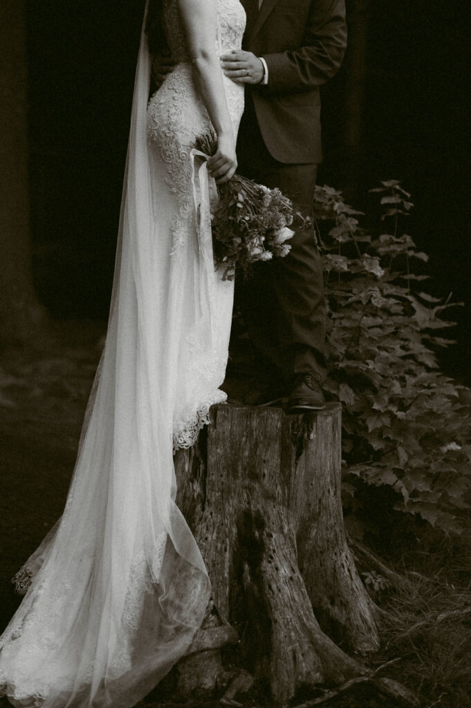 A moody black-and-white close-up of a bride and groom standing on a weathered tree stump. The groom holds the bride close as her lace gown cascades to the ground, bouquet in hand, with the forest fading into shadow behind them.