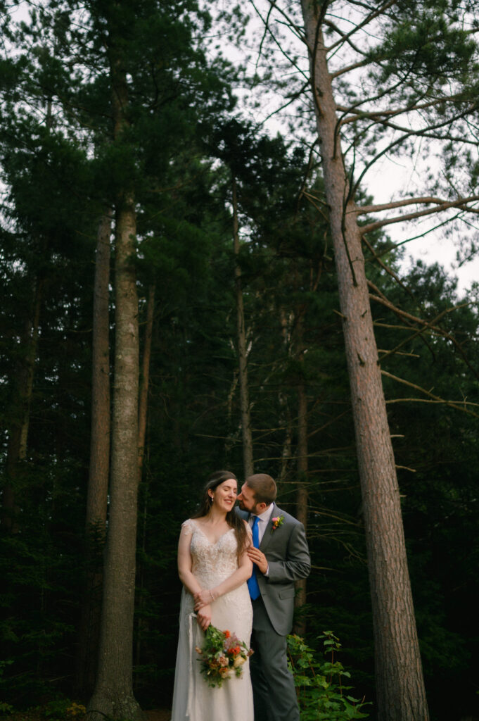 A groom leans in, grinning, as the bride tilts her head back in laughter. They stand among tall Adirondack pines, the last light of the day catching in the trees, making the moment feel both intimate and timeless.