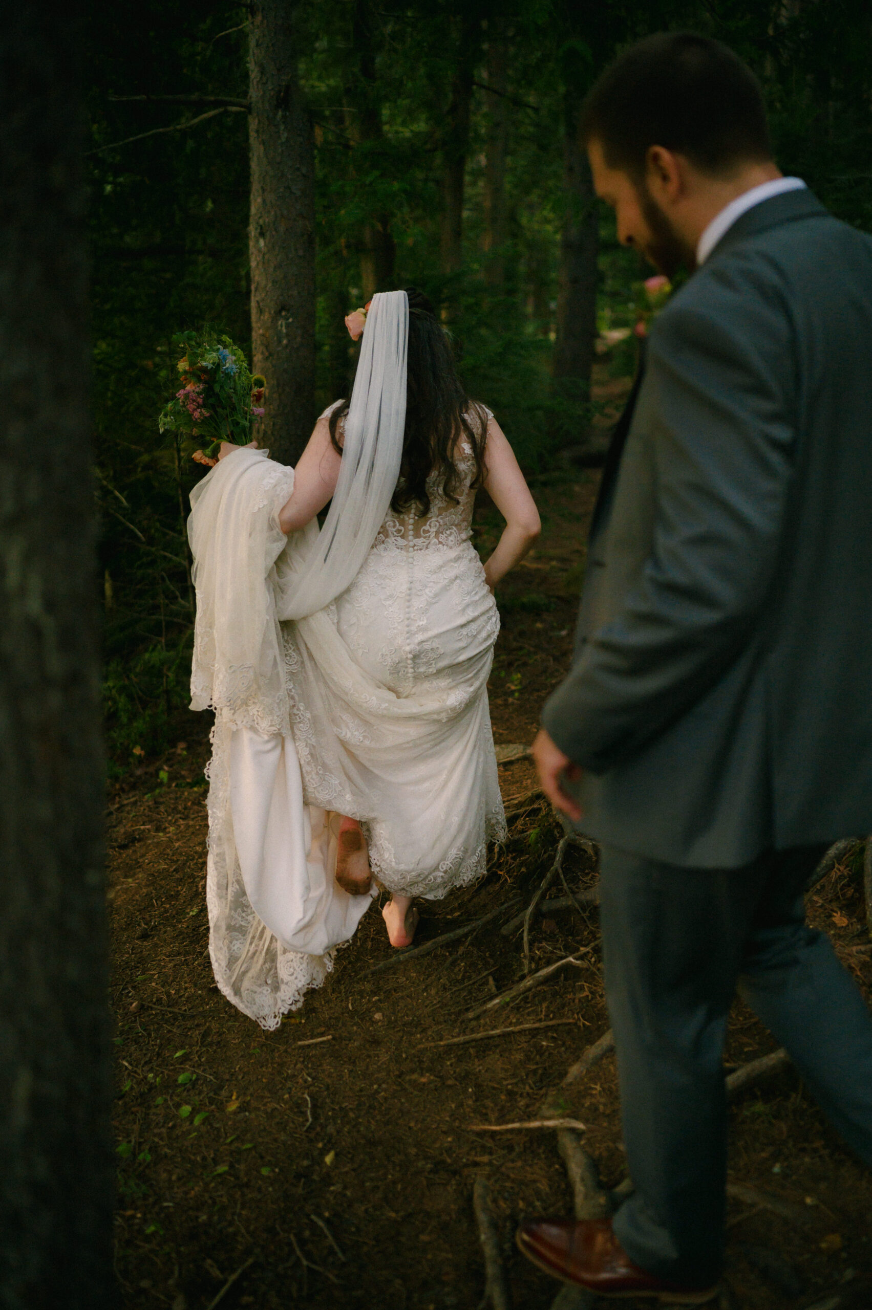 The bride, barefoot, lifts the train of her lace wedding gown as she walks through a wooded trail, holding her bouquet. The groom, dressed in a gray suit, follows closely behind, watching her navigate the path.