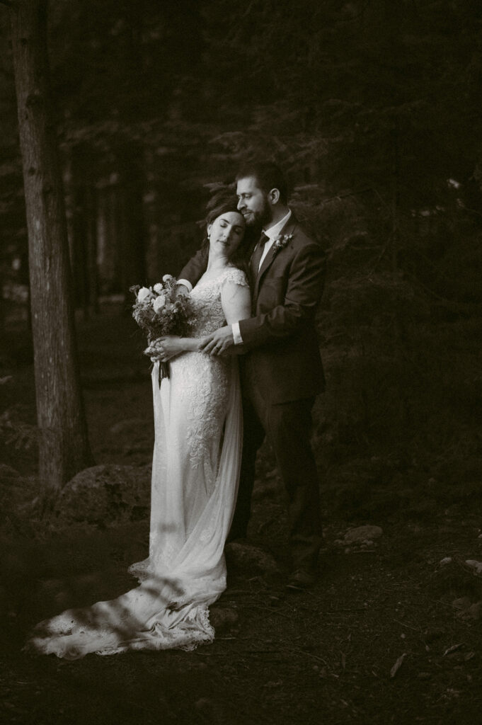 A black-and-white photo of a bride and groom standing in a forest. The bride leans into the groom’s embrace with her eyes closed, holding a bouquet of wildflowers. The groom gazes at her, his arms wrapped around her in an intimate moment.