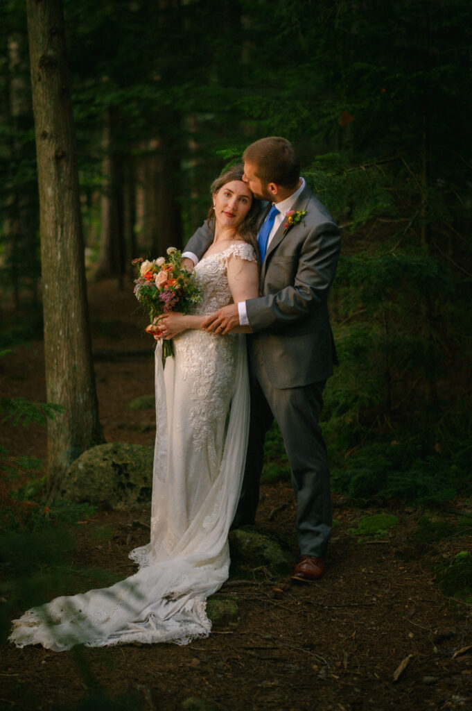 A bride and groom share a quiet moment in the forest. The bride holds a bouquet of flowers, looking softly into the distance, while the groom leans in to kiss her temple. Sunlight filters through the trees, casting a warm glow over them.