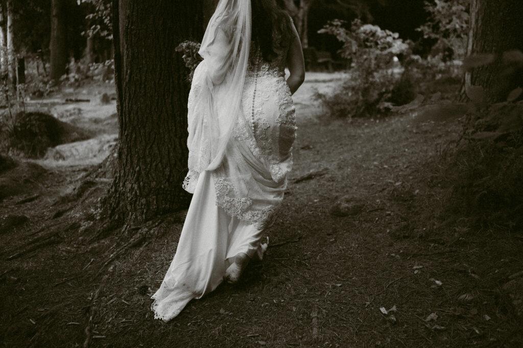 Black and white close-up of a bride walking through a wooded path. The back of her lace wedding dress and long veil trail behind her as she moves through the forest.