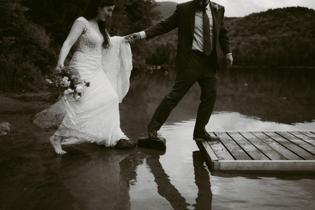 Candid black and white image of a newlywed couple stepping onto a dock. The bride, holding a bouquet, lifts her lace wedding dress as she carefully steps onto a rock, while the groom reaches out a hand to help her.