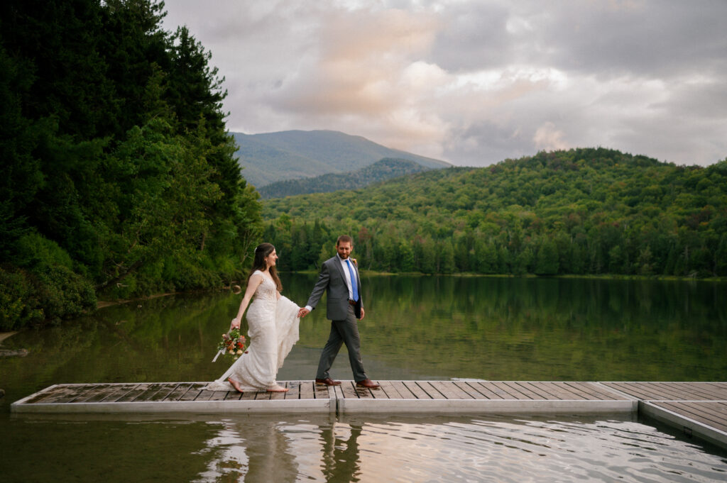 Bride and groom walking hand in hand along a floating dock. The serene mountain landscape reflects in the still water as they share a peaceful stroll at dusk.