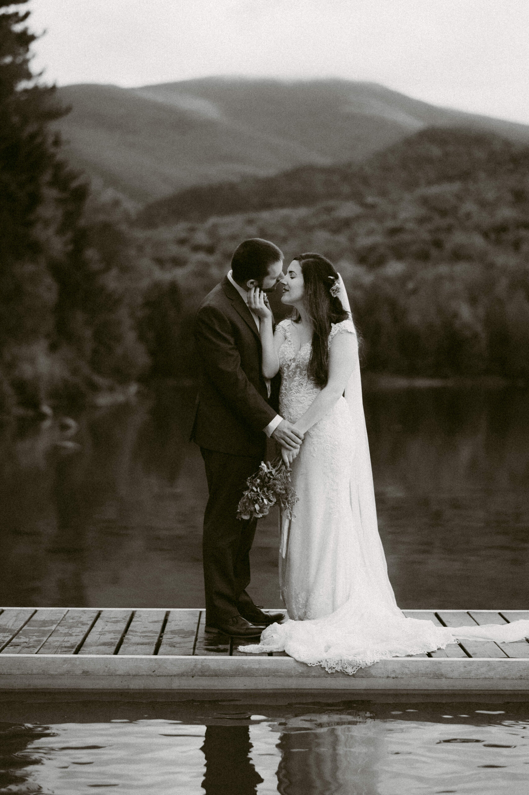 Black and white photo of a newlywed couple standing on a dock with mountain views. The groom tenderly cups the bride’s face as they lean in for a kiss, with misty mountains and a reflective lake in the background.