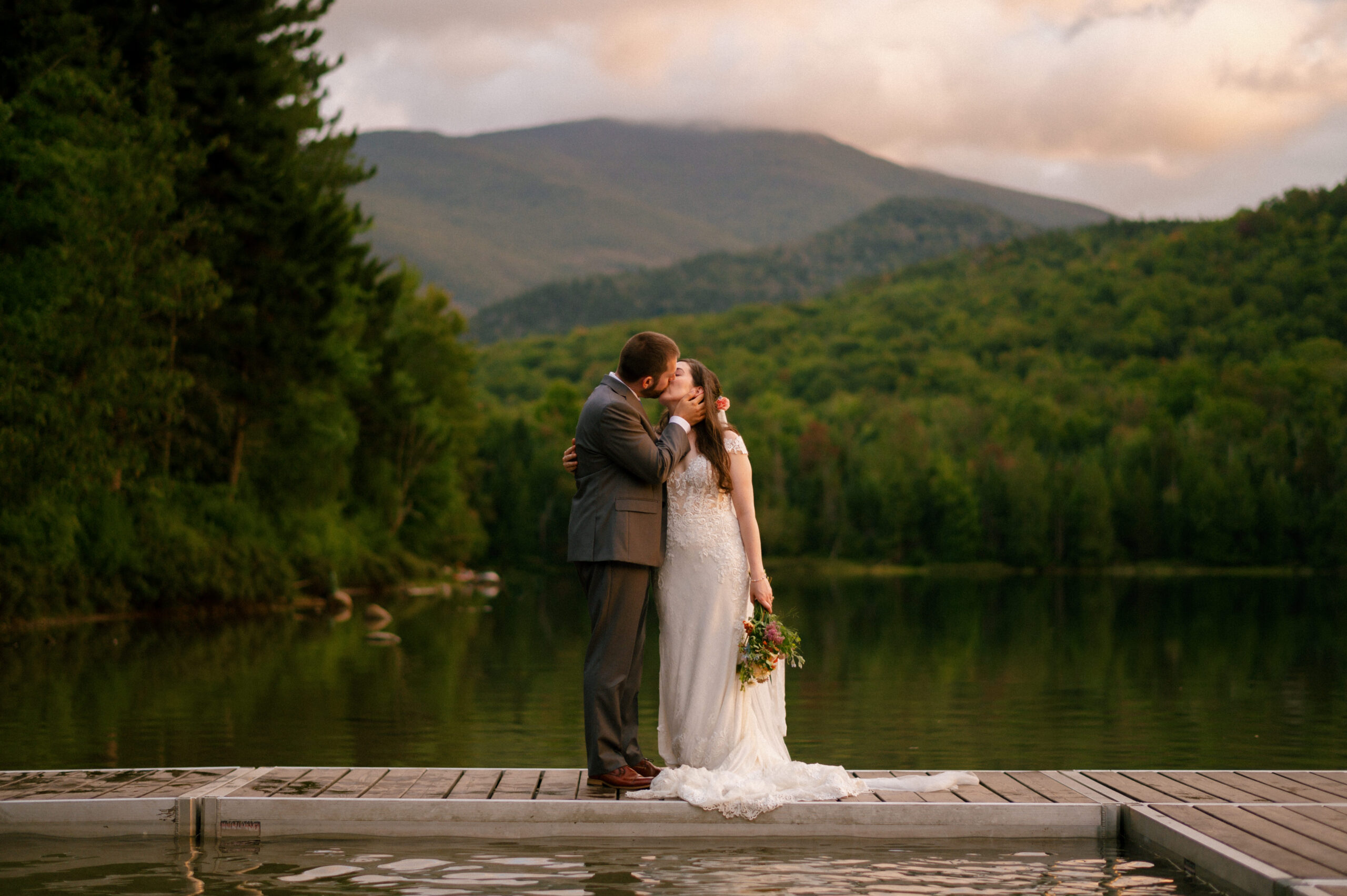 Romantic sunset kiss on the dock. A bride and groom embrace and kiss, bathed in golden light, with a peaceful lake and lush mountains behind them.