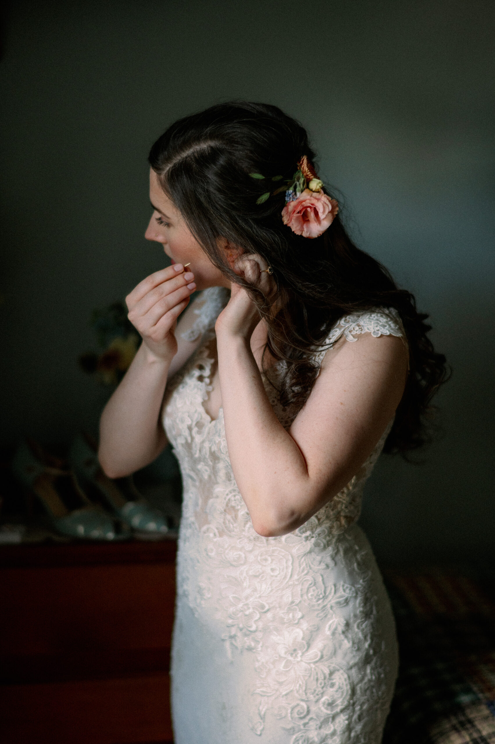 The bride turns slightly as she puts on her earring, her gaze focused downward. The intricate lace of her dress catches the light, highlighting its delicate texture. Her hairstyle, accented with fresh flowers, complements the vintage-inspired details of the room, creating a warm and personal getting-ready moment.