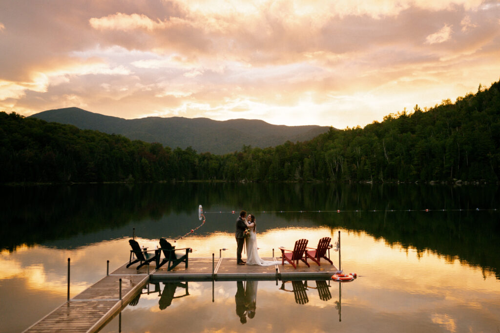 Wide-angle shot of a couple on a dock at sunset. Adirondack chairs and a lifebuoy frame the scene as the couple shares a quiet moment with a breathtaking sunset over the lake.