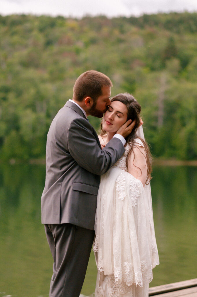 A groom in a gray suit gently kisses his bride’s forehead as she leans into his embrace, eyes closed, standing on a dock with a serene lake and lush green forest in the background.